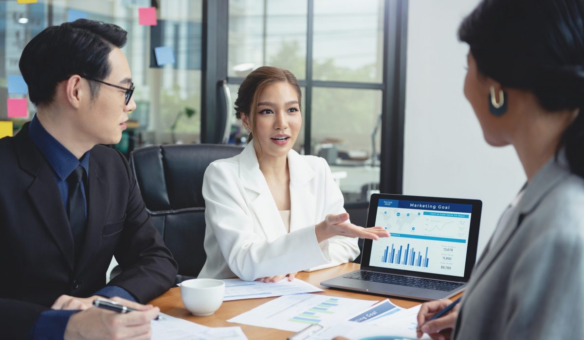 Group of Asian business professionals in a modern office setting discussing marketing goals and financial plans, with charts and graphs displayed on a laptop.